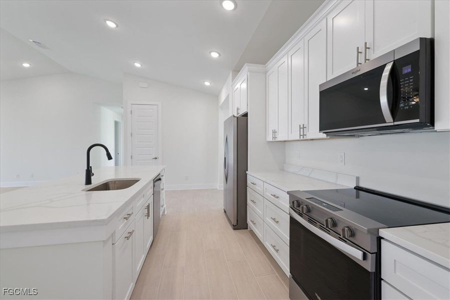 Kitchen with stainless steel appliances, light stone counters, white cabinetry, a center island with sink, and vaulted ceiling