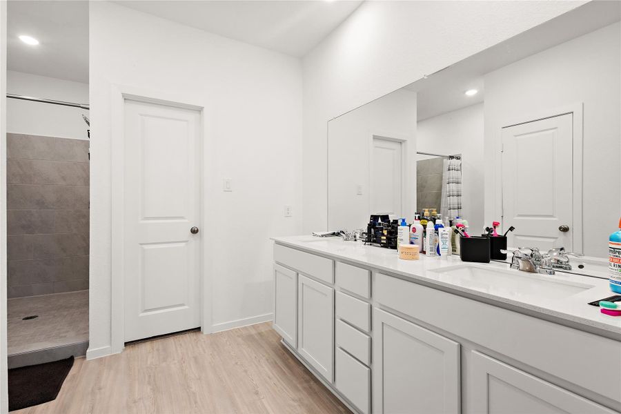 This bathroom features a modern design with a dual-sink vanity, ample counter space, and a large mirror. It has a walk-in shower with tiled walls and a sleek, light wood floor.