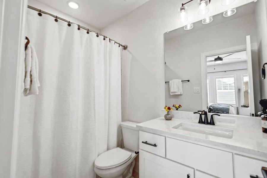 A clean, efficient bathroom is equipped with a crisp white vanity, quartz countertops, and a modern black faucet. The mirror reflects a glimpse of the connected bedroom and its windows, creating a bright atmosphere.