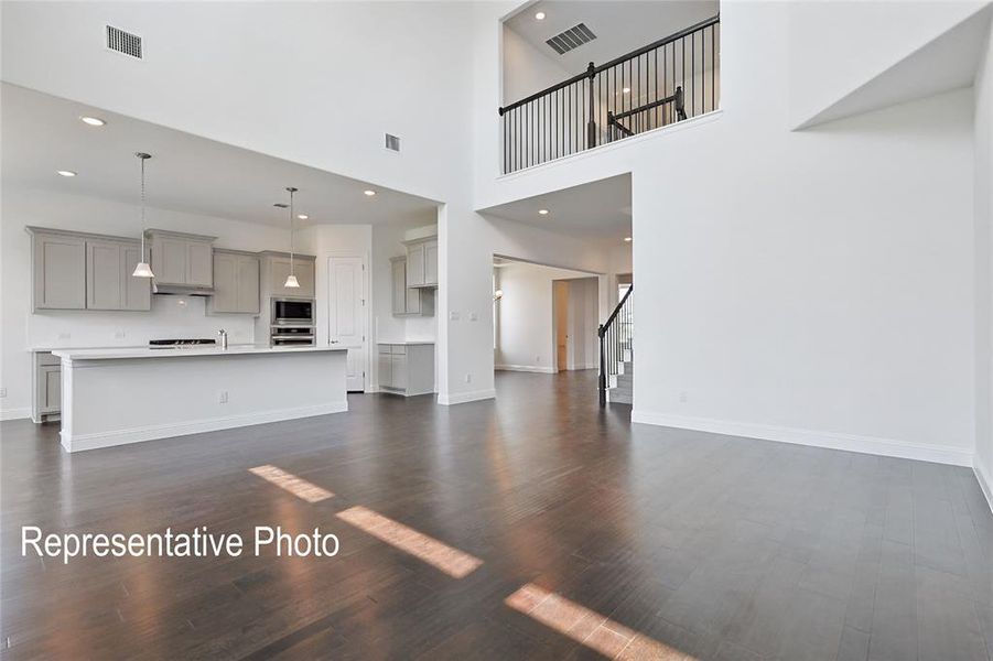 Expansive open-concept living space featuring high ceilings, wood-finish flooring, a kitchen island with pendant lighting, and a second-story landing with wrought iron balusters