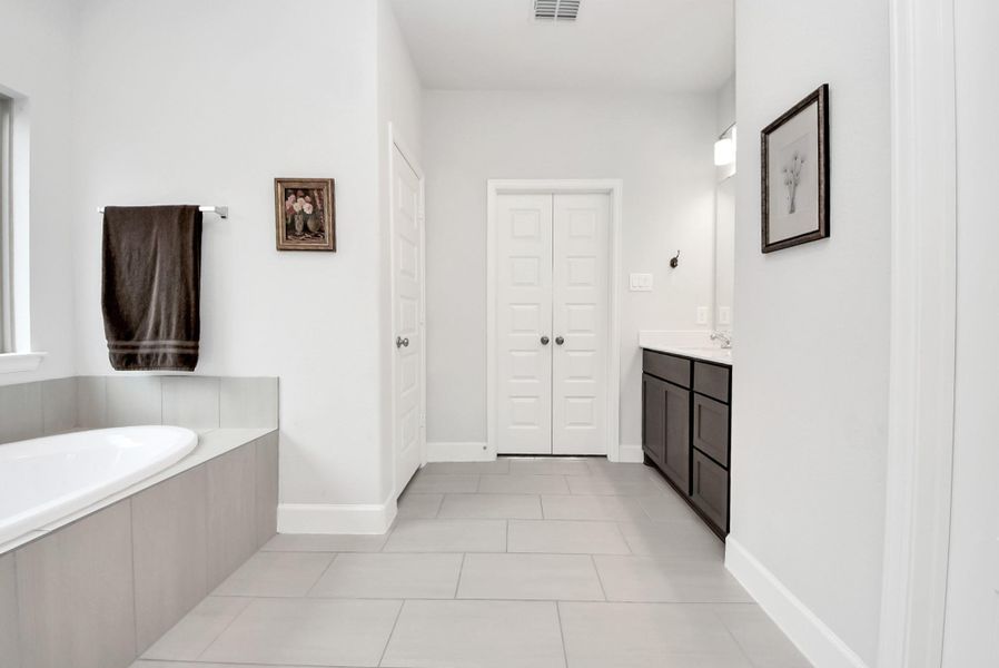A modern bathroom featuring a large soaking bathtub, sleek dark cabinetry, and light-colored tiles. Minimalist decor and a double-door closet create a clean, refined space with a serene atmosphere.