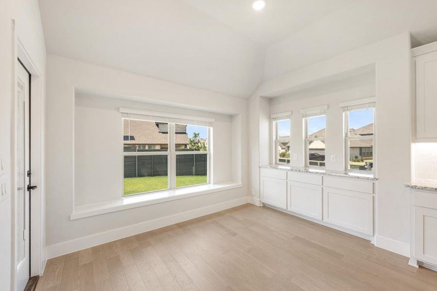 Bright empty dining area with light wood floors, white walls, large windows, and built-in white cabinetry with stone countertop.