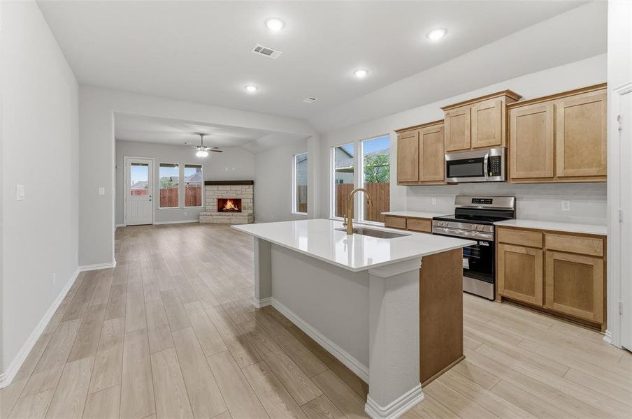 Spacious open-concept kitchen and living area featuring wood-finish flooring, a contemporary kitchen island with a single-basin sink, light wood cabinetry, stainless steel appliances, and a stone fireplace