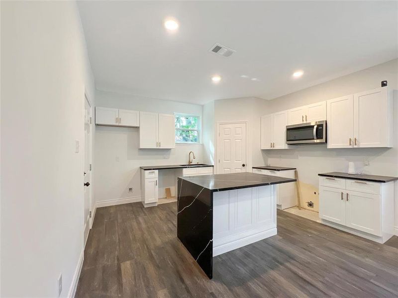 Kitchen featuring dark wood finished floors, white cabinetry, stainless steel microwave, dark countertops, and a center island Kitchen featuring dark wood finished floors, white cabinetry, stainless steel microwave, dark countertops, and a center island