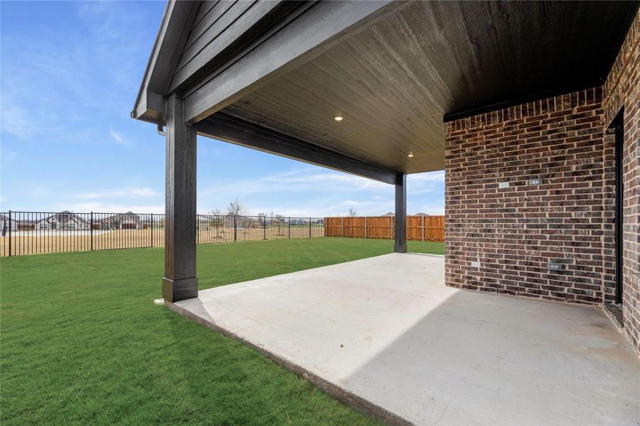 Covered back patio with concrete slab, green lawn, and fenced backyard next to brick home exterior Covered back patio with concrete slab, green lawn, and fenced backyard next to brick home exterior