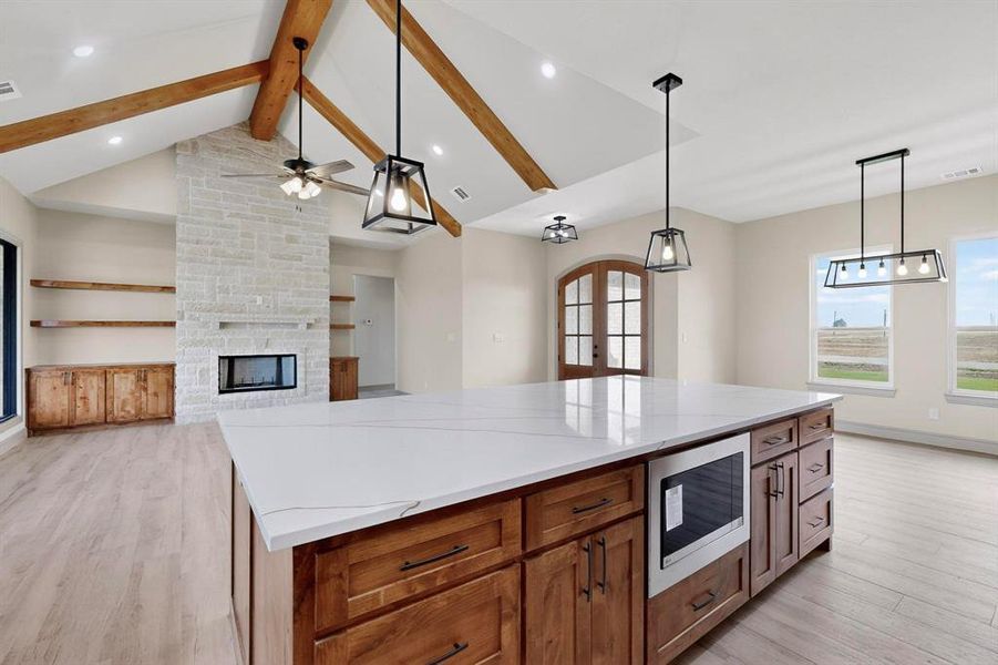 Kitchen featuring brown cabinetry, pendant lighting, stainless steel microwave, beamed ceiling, and a stone fireplace Kitchen featuring brown cabinetry, pendant lighting, stainless steel microwave, beamed ceiling, and a stone fireplace