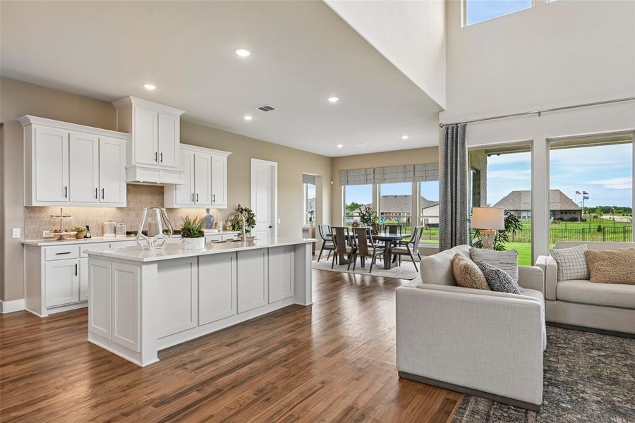 Open-concept kitchen and living area with white cabinets, large island, hardwood floors, and dining space near large windows Open-concept kitchen and living area with white cabinets, large island, hardwood floors, and dining space near large windows