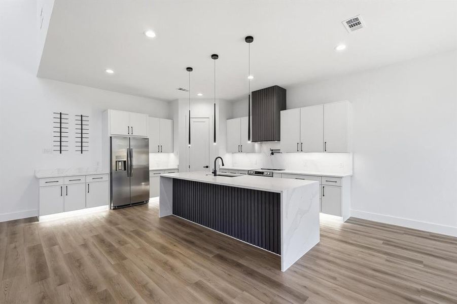 Kitchen featuring hanging light fixtures, a kitchen island with sink, stainless steel fridge, white cabinets, and light stone counters Kitchen featuring hanging light fixtures, a kitchen island with sink, stainless steel fridge, white cabinets, and light stone counters