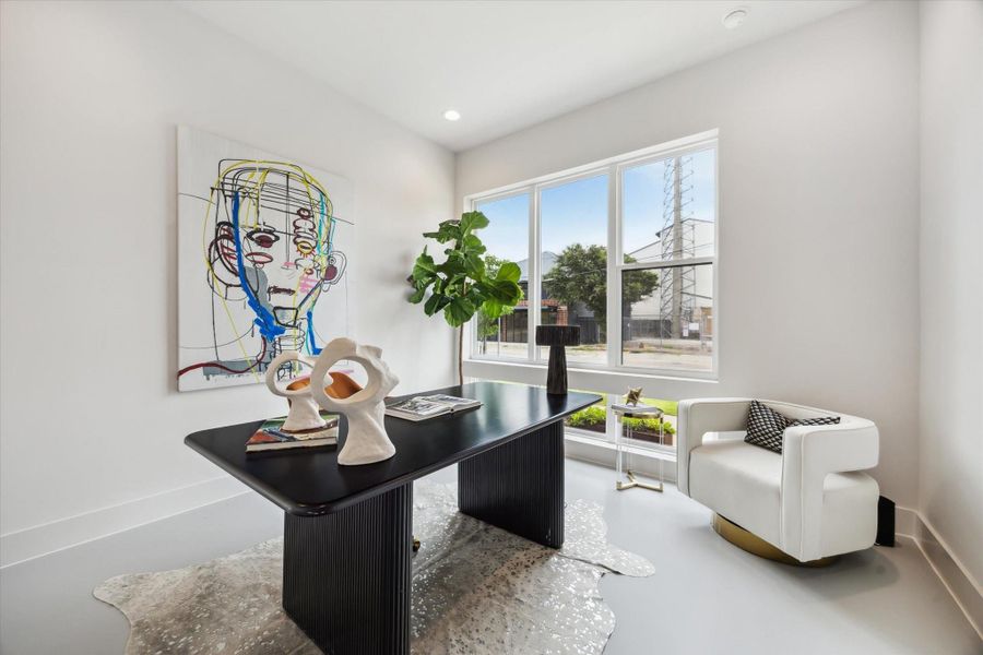 This bright, modern home office features a sleek black desk, contemporary art, and a large window letting in natural light. A stylish white armchair and decorative accents add sophistication, while a lush plant brings a touch of nature indoors.