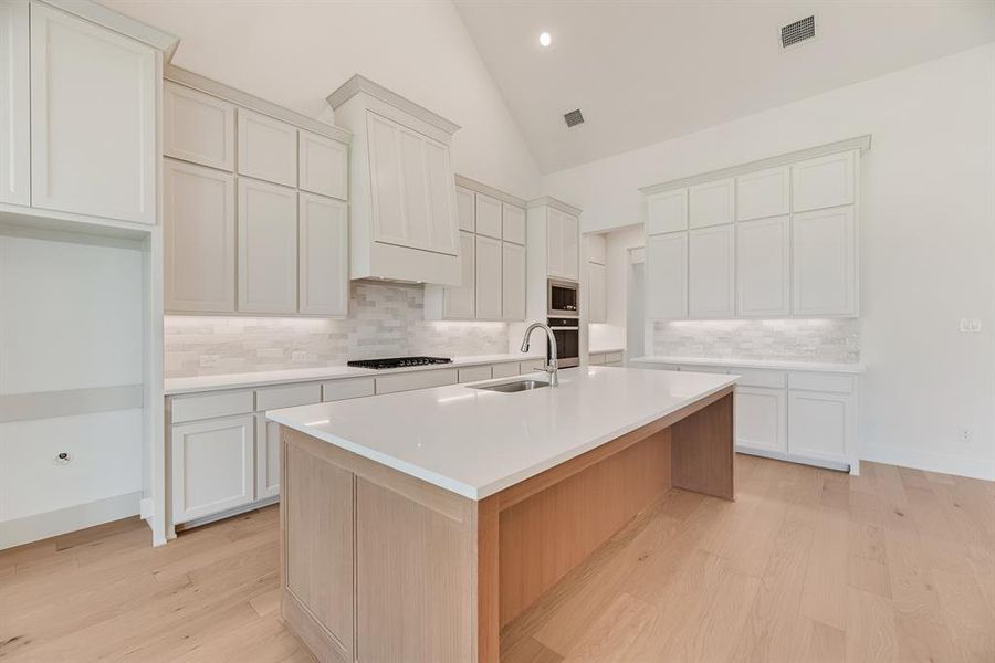 Kitchen featuring backsplash, light wood-style floors, a kitchen island with sink, high vaulted ceiling, and light stone counters