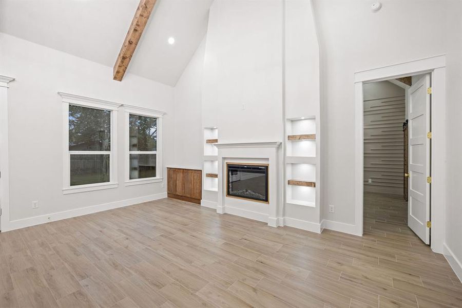 Spacious room featuring vaulted ceilings with an exposed beam, wood-finish flooring, and an electric fireplace with built-in shelving Spacious room featuring vaulted ceilings with an exposed beam, wood-finish flooring, and an electric fireplace with built-in shelving