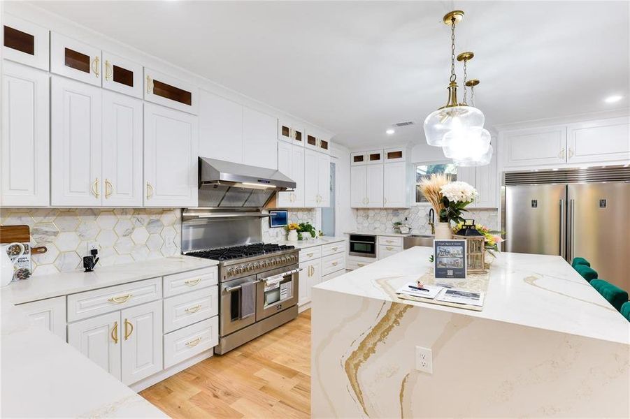 Kitchen with hanging light fixtures, high end appliances, white cabinets, and wall chimney exhaust hood