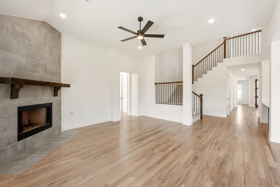 Open living room with tile fireplace, wood mantel, hardwood floors, ceiling fan, and staircase with iron railings