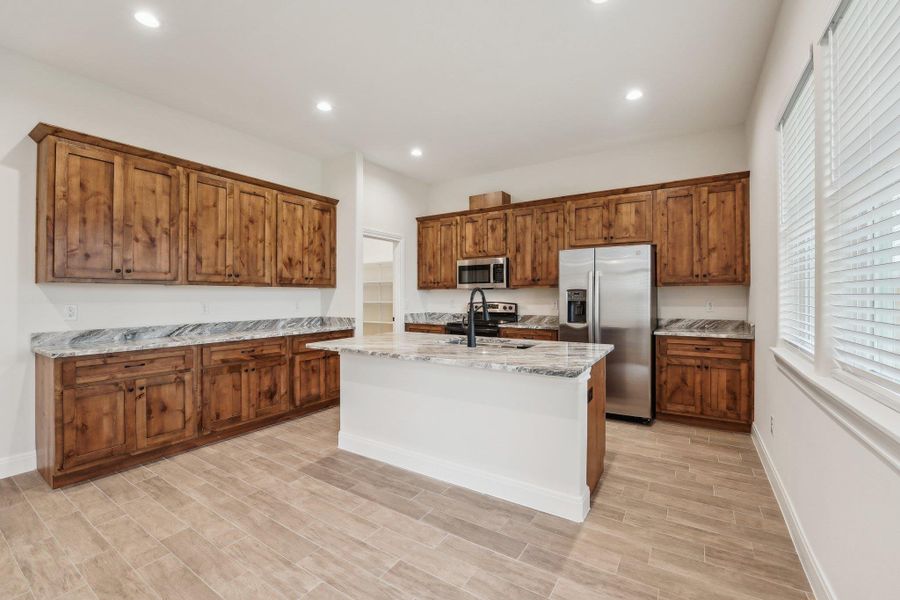 Kitchen featuring light stone counters, a center island with sink, a sink, appliances with stainless steel finishes, and brown cabinets Kitchen featuring light stone counters, a center island with sink, a sink, appliances with stainless steel finishes, and brown cabinets