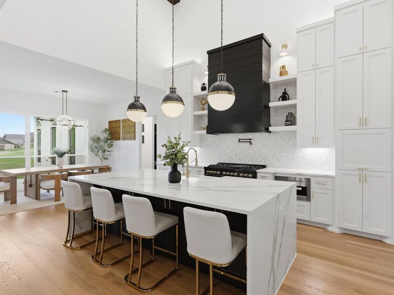 Expansive kitchen featuring a large central island with waterfall countertop, integrated cooktop with custom range hood, full-height white cabinetry with gold-tone hardware, and light wood-finish flooring