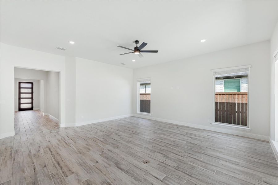 Open living room with light wood-look floors, ceiling fan, two windows, and view to front entry door