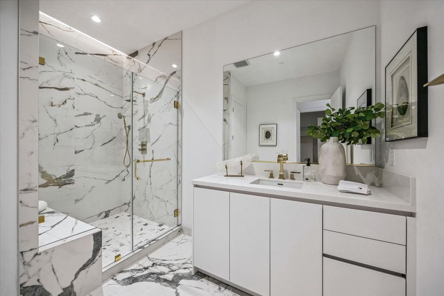 The second primary bath is beautifully appointed with sleek, modern finishes, featuring a spacious glass-enclosed shower wrapped in striking marble, complemented by elegant gold fixtures. A clean-lined vanity with storage, designer lighting & a large mirror create a bright, spa-like setting that blends style & functionality.