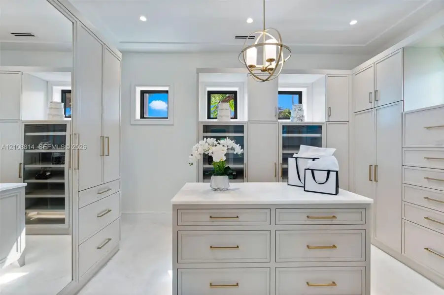 Custom cabinetry featuring glass-front display doors and integrated shelving, central storage island with stone-finish countertop, brushed brass hardware, mirrored door panels, and spherical pendant lighting Custom cabinetry featuring glass-front display doors and integrated shelving, central storage island with stone-finish countertop, brushed brass hardware, mirrored door panels, and spherical pendant lighting