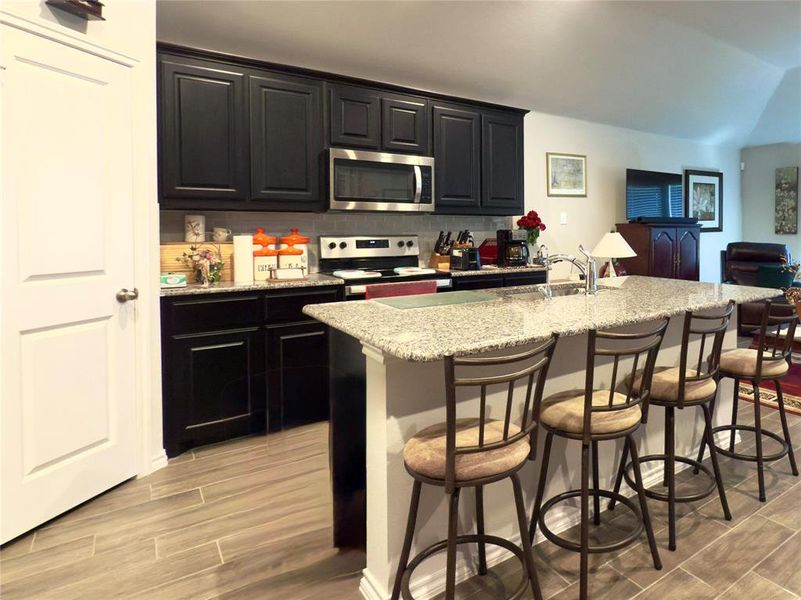 Kitchen with backsplash, wood tiled floors, light stone counters, stainless steel appliances, and dark cabinetry Kitchen with backsplash, wood tiled floors, light stone counters, stainless steel appliances, and dark cabinetry