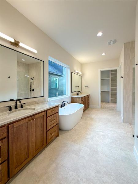 Spacious bathroom featuring twin wood-finish vanities with light stone countertops, a freestanding white soaking tub, and large format floor tiling
