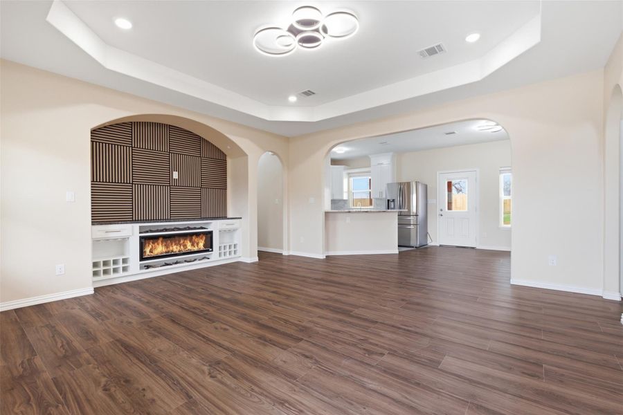 Unfurnished living room with arched walkways, dark wood-style floors, a raised ceiling, a glass covered fireplace, and recessed lighting