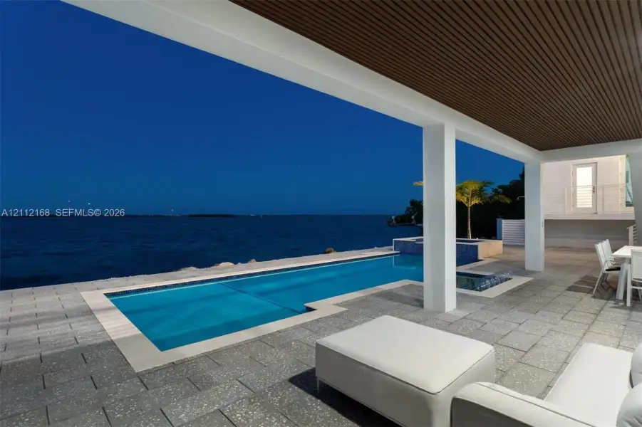 Covered patio with wood-slat ceiling, natural stone tile flooring, swimming pool, built-in spa, and water frontage Covered patio with wood-slat ceiling, natural stone tile flooring, swimming pool, built-in spa, and water frontage