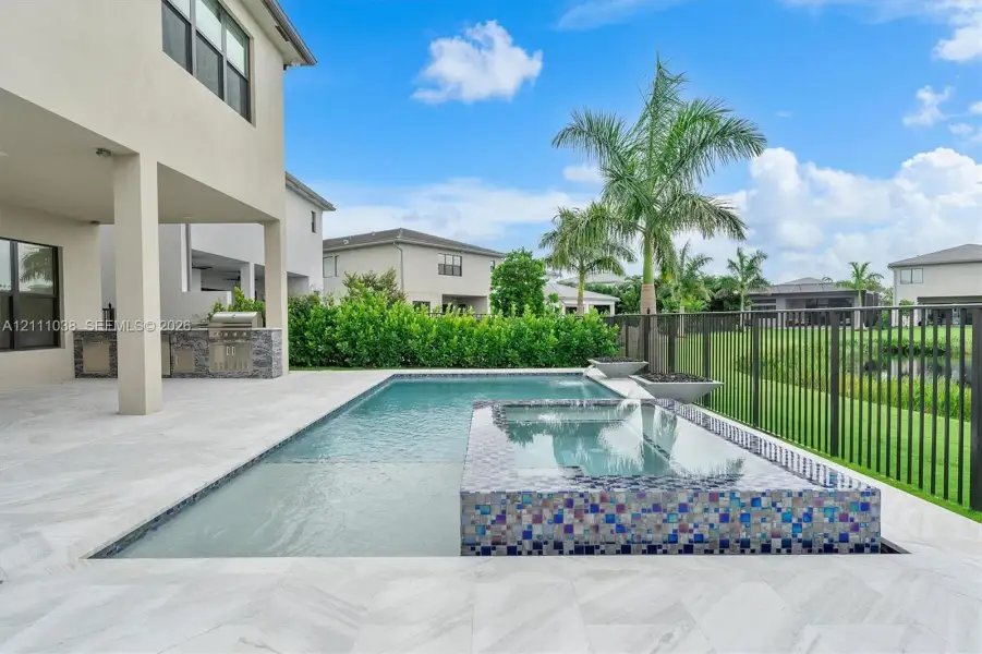 Covered patio featuring a built-in outdoor kitchen, stone-tile pool deck, swimming pool with integrated spillover spa, and ornamental metal perimeter fencing