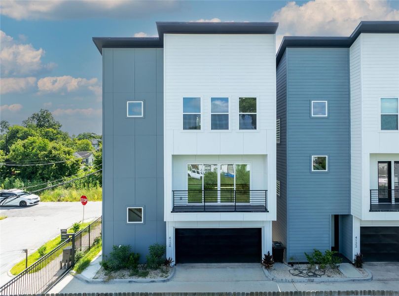 Modern three-story townhouse with a sleek exterior featuring a combination of gray and white paneling. Large windows provide ample natural light, and the property includes a gated entrance with a garage. Located on a corner lot with a view of greenery.