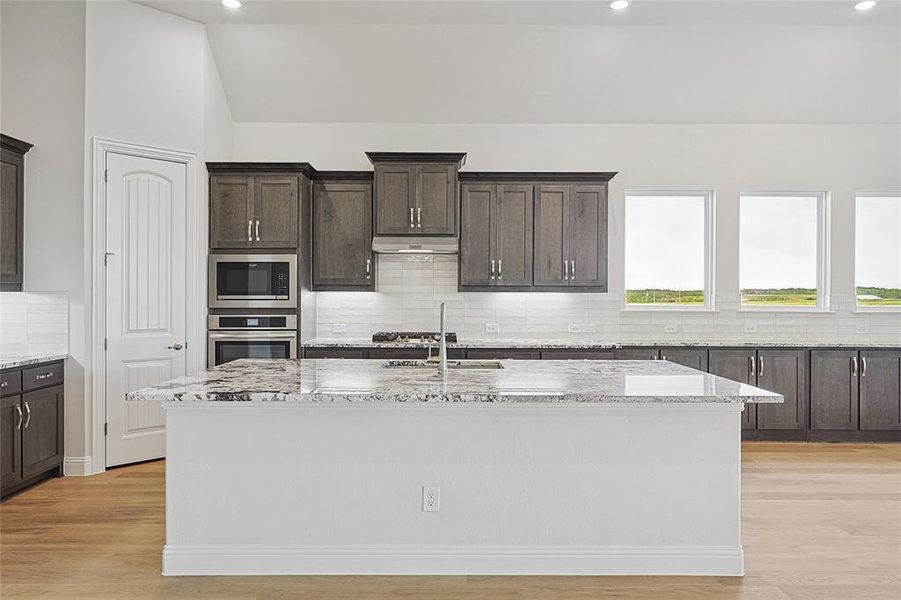 Kitchen with dark wood finish cabinetry, light stone countertops, backsplash, light wood-style flooring, and an island with sink