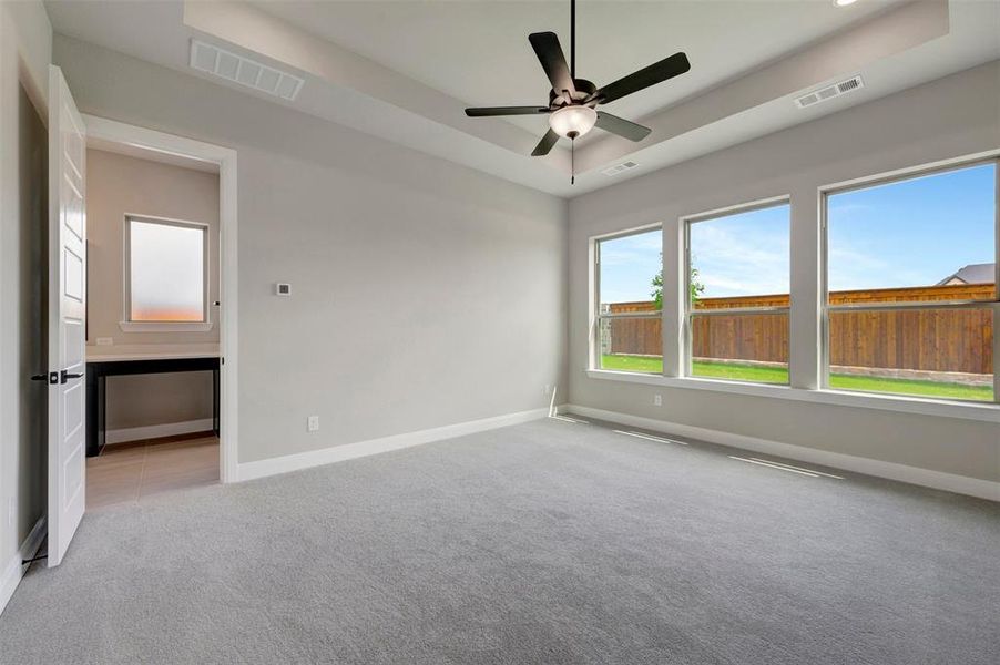 Spacious room featuring light gray carpet flooring, a tray ceiling with a ceiling fan, and three large windows overlooking the backyard with a privacy fence