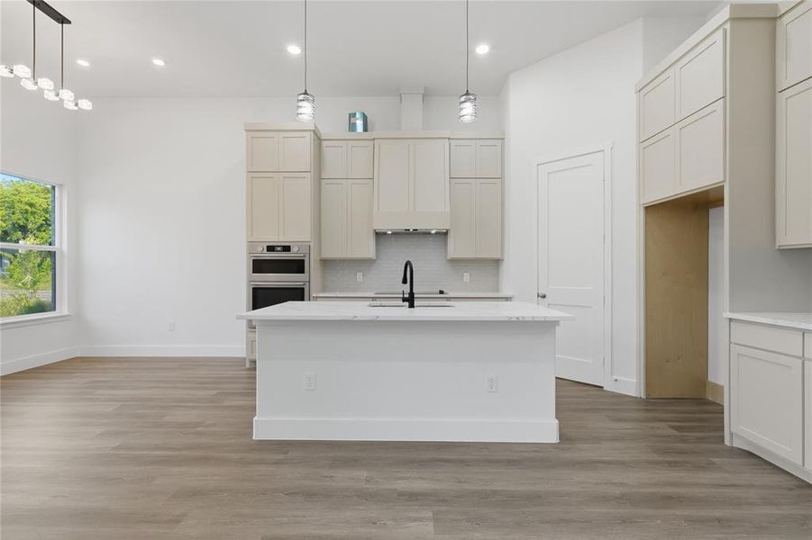 Modern kitchen featuring an expansive island with a sink, light-toned cabinetry, built-in double ovens, a subway tile backsplash, and wood-finish flooring
