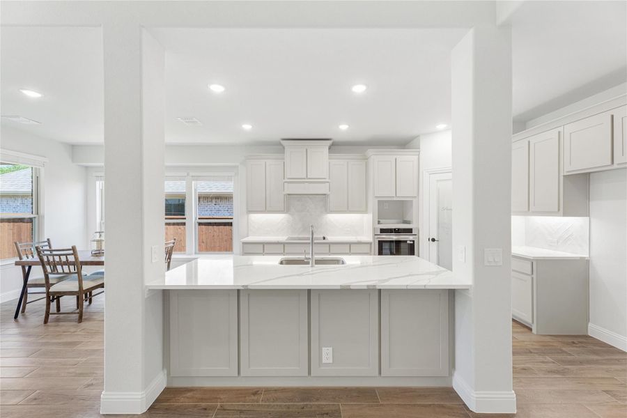 Open kitchen with large white quartz island, gray cabinets, herringbone backsplash, and wood-look tile floors Open kitchen with large white quartz island, gray cabinets, herringbone backsplash, and wood-look tile floors