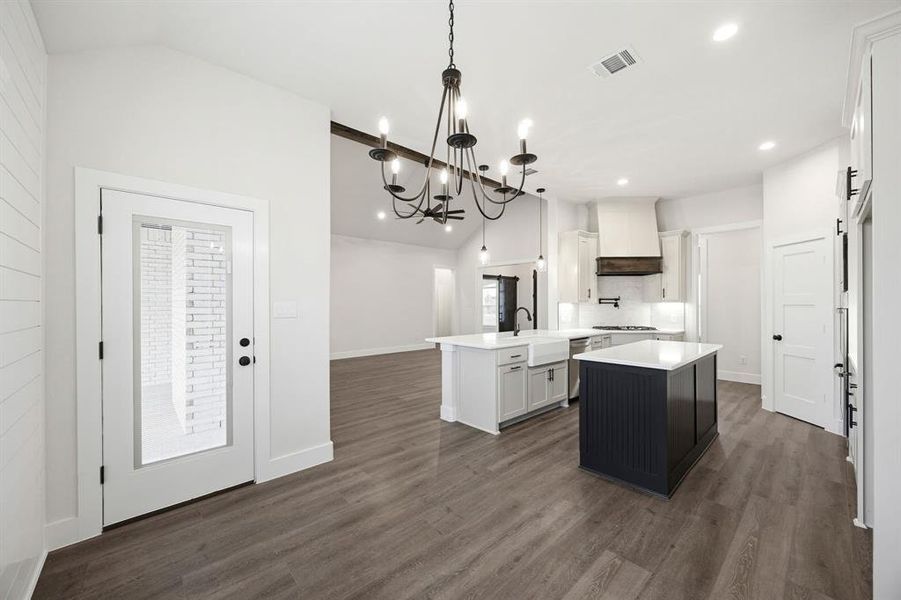 Open-concept kitchen and living area featuring wood-finish flooring, two kitchen islands with solid surface countertops, custom range hood, and a black wrought iron chandelier