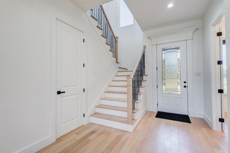 Entryway featuring light wood-finish flooring, a staircase with black metal balusters and wood-finish handrail, white paneled doors with black hardware, a recessed ceiling light, and a white front door with decorative glass inserts Entryway featuring light wood-finish flooring, a staircase with black metal balusters and wood-finish handrail, white paneled doors with black hardware, a recessed ceiling light, and a white front door with decorative glass inserts