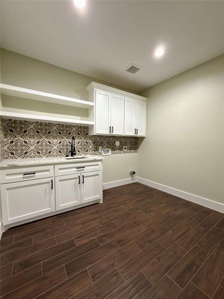 This photo showcases a modern laundry room with white cabinetry, open shelving, and a stylish patterned backsplash. The space has a dark wood-look tile floor, recessed lighting, and a utility sink, offering both functionality and a contemporary design. This photo showcases a modern laundry room with white cabinetry, open shelving, and a stylish patterned backsplash. The space has a dark wood-look tile floor, recessed lighting, and a utility sink, offering both functionality and a contemporary design.