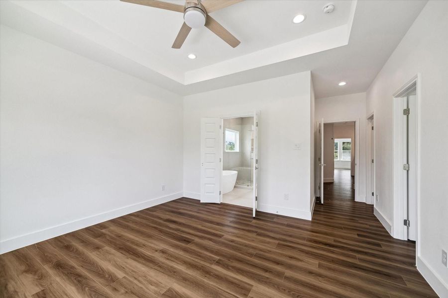 An alternate view of the primary suite showcases its impressive proportions, tray ceiling, contemporary ceiling fan, recessed lighting, and seamless wood-look flooring. Double doors open directly to the private primary bathroom, creating a convenient suite-style layout. The bright neutral palette enhances the sense of space and offers an elegant foundation for any décor. Multiple doorways provide easy access to adjoining areas while preserving the room’s open and comfortable feel. An alternate view of the primary suite showcases its impressive proportions, tray ceiling, contemporary ceiling fan, recessed lighting, and seamless wood-look flooring. Double doors open directly to the private primary bathroom, creating a convenient suite-style layout. The bright neutral palette enhances the sense of space and offers an elegant foundation for any décor. Multiple doorways provide easy access to adjoining areas while preserving the room’s open and comfortable feel.