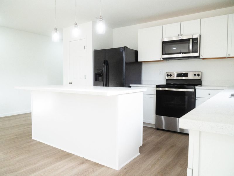 Modern kitchen with clean, white cabinetry and countertops. Features stainless steel appliances, a large black fridge, and a central island with pendant lighting. Bright and spacious with light wood flooring.
