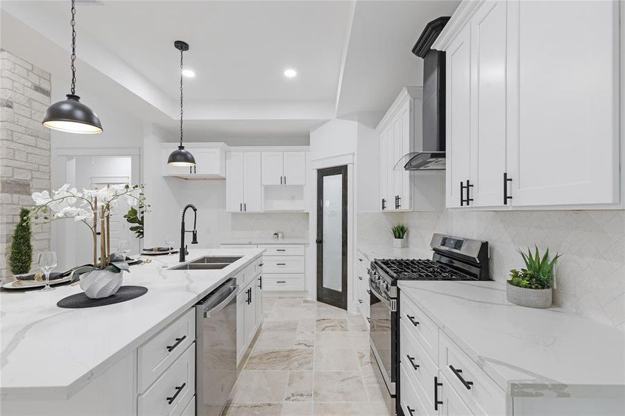 This is a modern kitchen with white cabinetry, stainless steel appliances, a gas range, and an island with pendant lighting. The space features stone flooring and a clean, bright color palette.