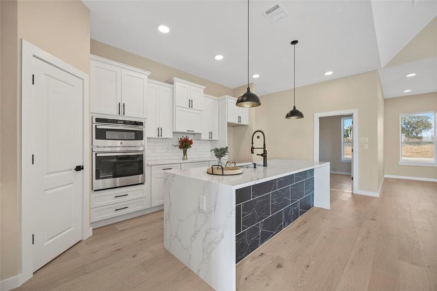 Kitchen with double oven, a center island with sink, decorative light fixtures, light wood-type flooring, and white cabinets