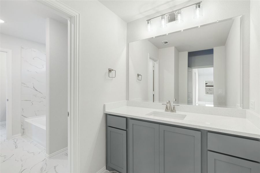 Bathroom with gray vanity, white quartz countertop, large mirror, and marble tile floors with soaking tub visible in background.