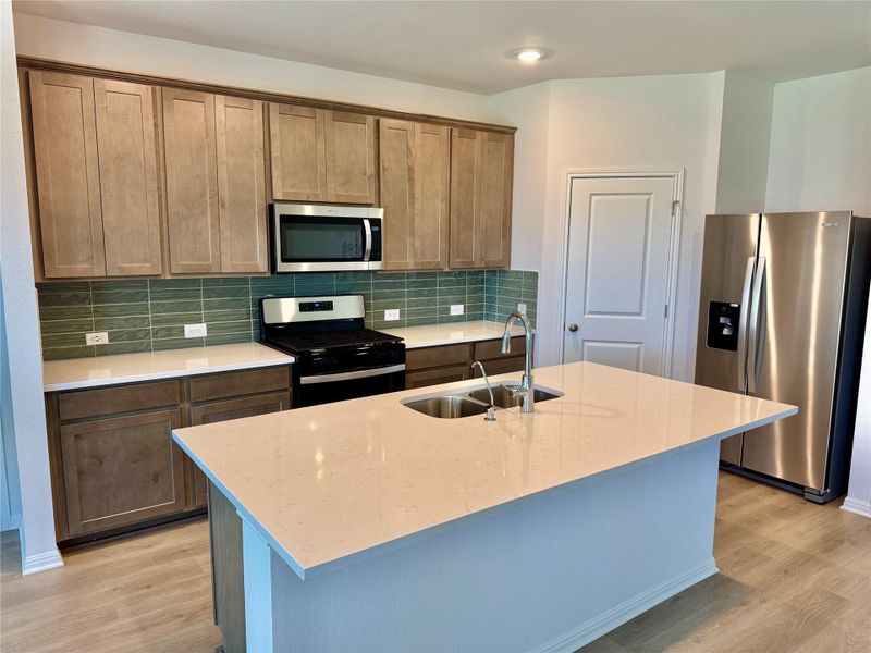 Kitchen with appliances with stainless steel finishes, decorative backsplash, light wood finished floors, an island with sink, and light stone counters