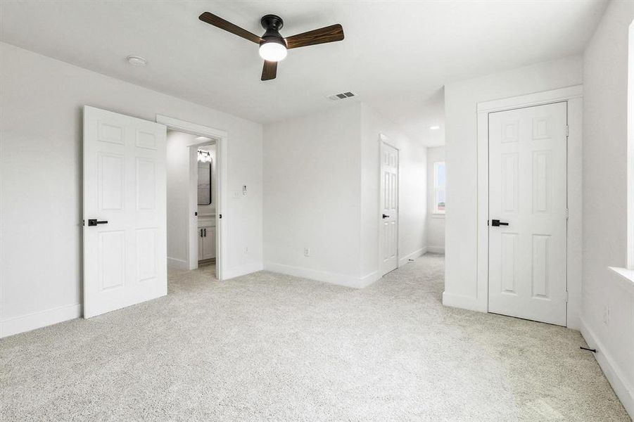 Carpeted room with a ceiling fan, featuring multiple white six-panel doors with black hardware, and a partial view into a bathroom with a vanity and mirror Carpeted room with a ceiling fan, featuring multiple white six-panel doors with black hardware, and a partial view into a bathroom with a vanity and mirror