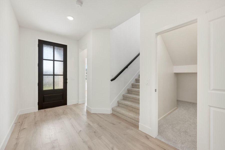 Bright entryway with black glass-panel front door, light hardwood floors, and carpeted staircase with dark handrail