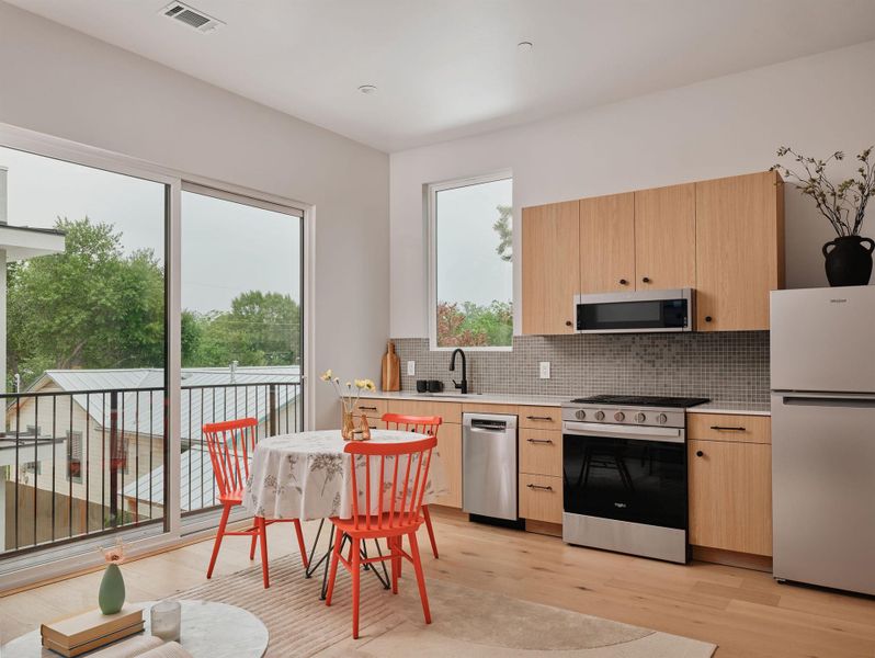 Light wood-finish flooring complements the contemporary kitchen featuring wood-tone cabinetry, stainless steel appliances, and a mosaic tile backsplash