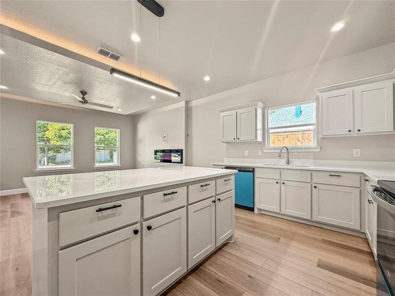 Kitchen featuring a large central island with a solid surface countertop, white shaker-style cabinetry, and wood-finish flooring Kitchen featuring a large central island with a solid surface countertop, white shaker-style cabinetry, and wood-finish flooring