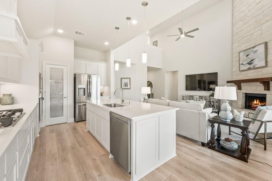 Open-concept kitchen with white island and stainless appliances flowing into living room with stone fireplace and high ceilings Open-concept kitchen with white island and stainless appliances flowing into living room with stone fireplace and high ceilings
