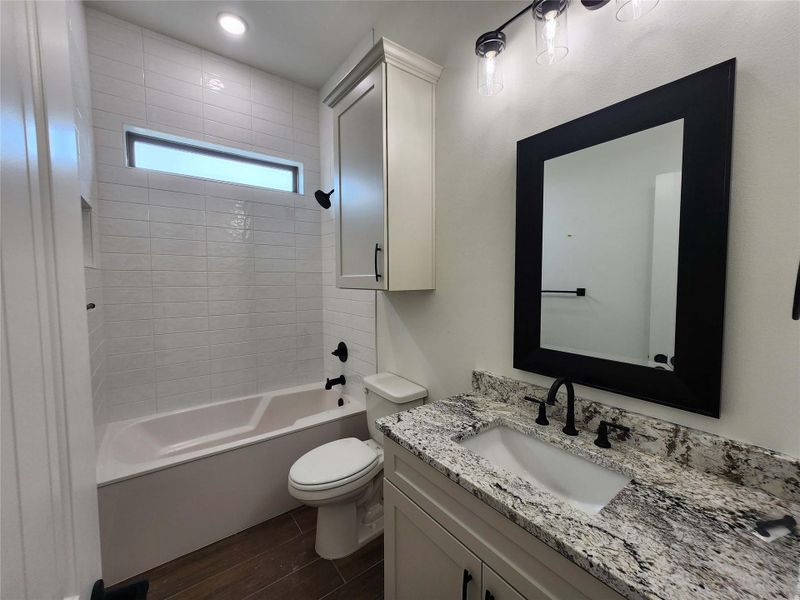 Bathroom featuring an undermount sink with a granite countertop, matte black fixtures, a framed mirror, a tiled shower-tub combination, and wood-finish flooring