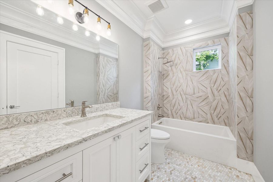 This secondary bathroom features a modern design with a large vanity and marble countertop, a sleek bathtub with decorative tiled walls, and a bright atmosphere enhanced by recessed lighting and a small window.