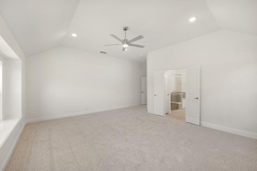 Empty bedroom with vaulted ceiling, ceiling fan, carpet flooring, and open ensuite bathroom door