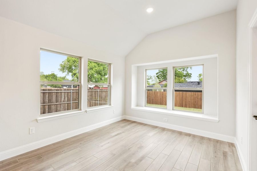 Empty room with vaulted ceiling, light wood floors, and large windows overlooking a fenced backyard